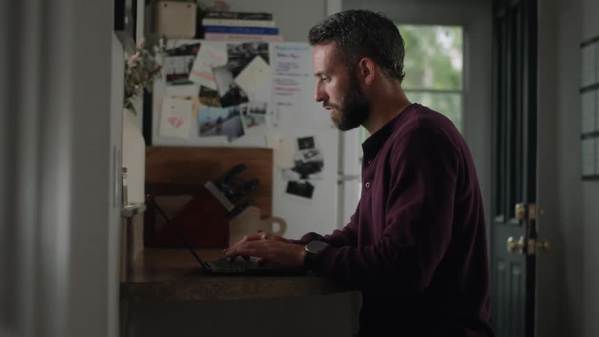A bearded man sits at a small desk working on a laptop in a room handling a smartphone