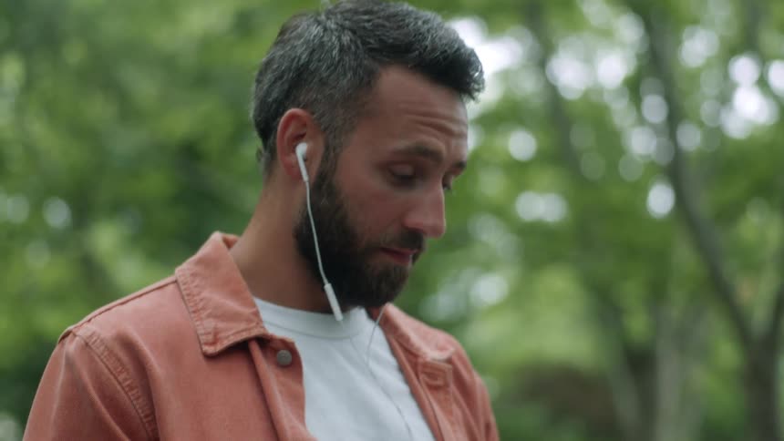 Man sitting in a park using a laptop and headphones while taking a phone call