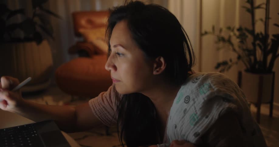Woman sitting on the floor holding her baby on her lap in a living room and working on a tablet with a digital pen