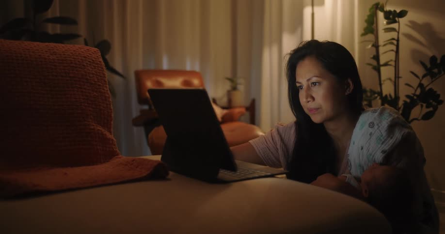Mother holding her baby while sitting on the floor in the living room uses a stylus on atouchscreen