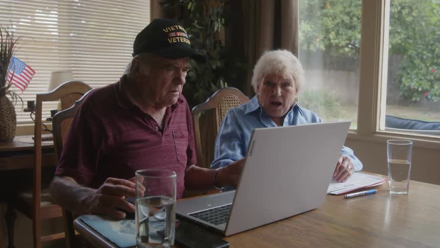 Two elderly people sitting at a table with documents and a laptop