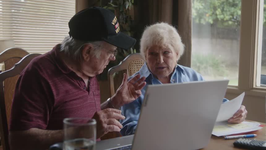 Elderly couple sitting at a table looking at documents and using a laptop