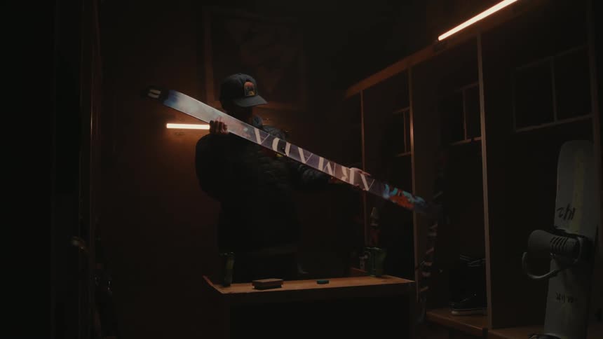 A man wearing a cap and a coat visually inspects a snow ski in a dingy room