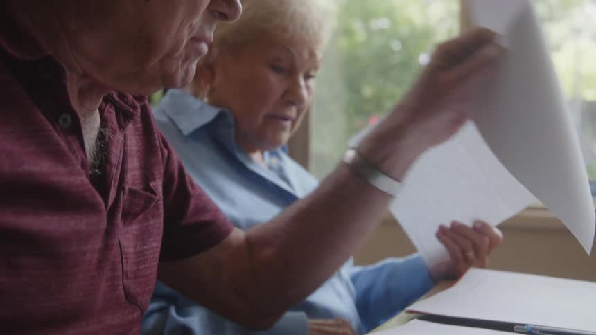 An elderly couple sitting at home checking documents and papers near a window
