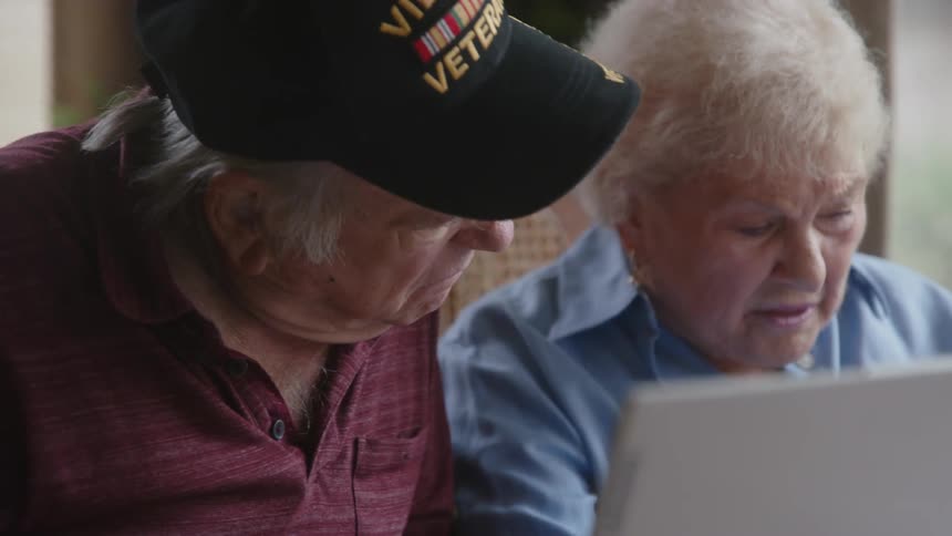 Two elderly people sitting at a table using a laptop and talking
