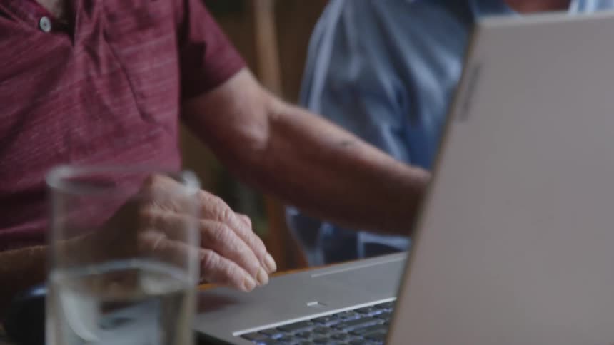 An old man sitting next to an old woman reaches out and pushes a laptop button