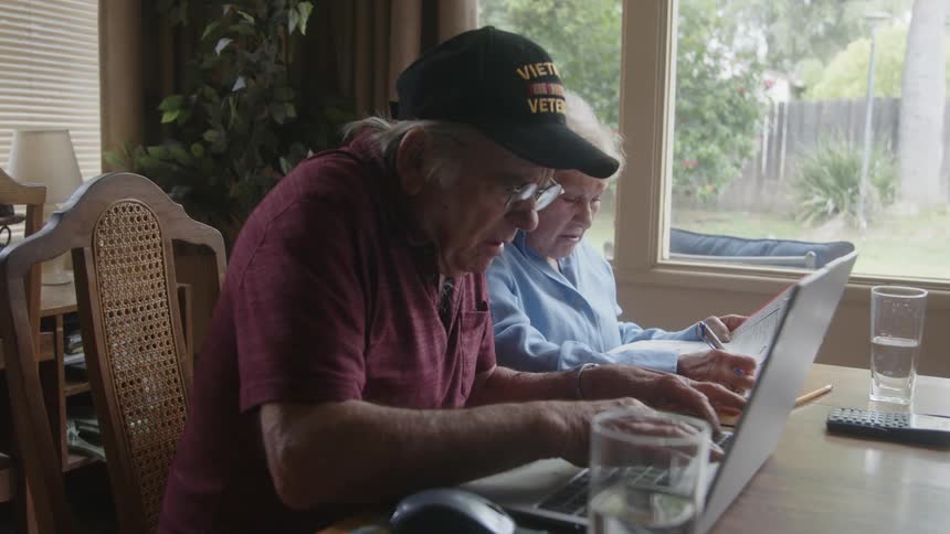 Two elderly people sitting at a table analyzing documents and using a laptop talking to each other