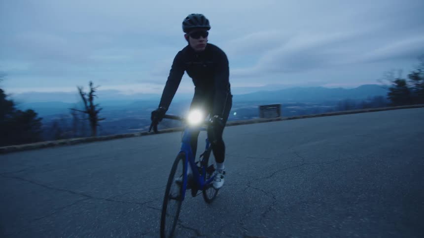 A bicyclist wearing sunglasses at dusk rides a bike on a rural road