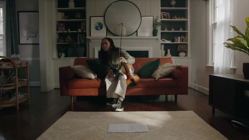 An asian woman holding a violin walks over to a couch and sits down