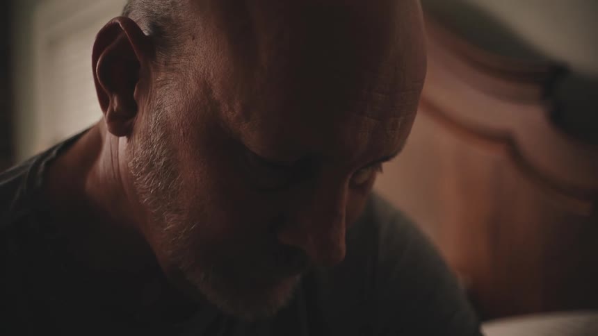 An older white man stands in a bedroom with his head down looking sad