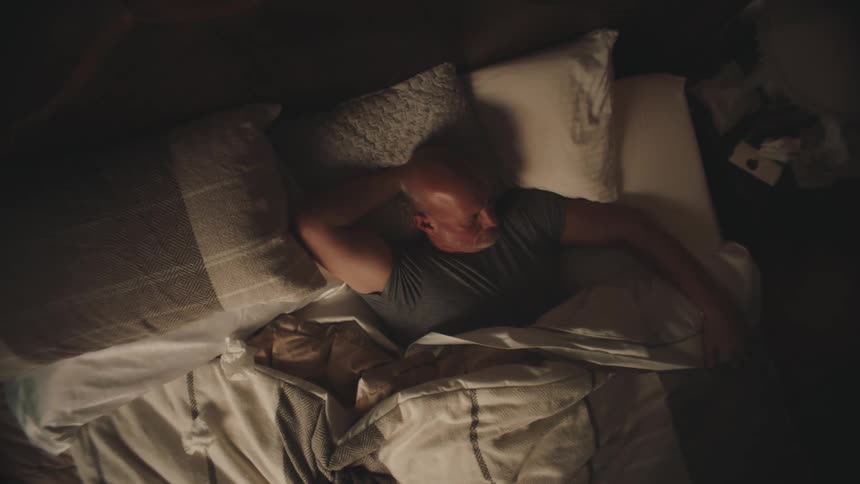 A balding man moves restlessly in a cluttered bed