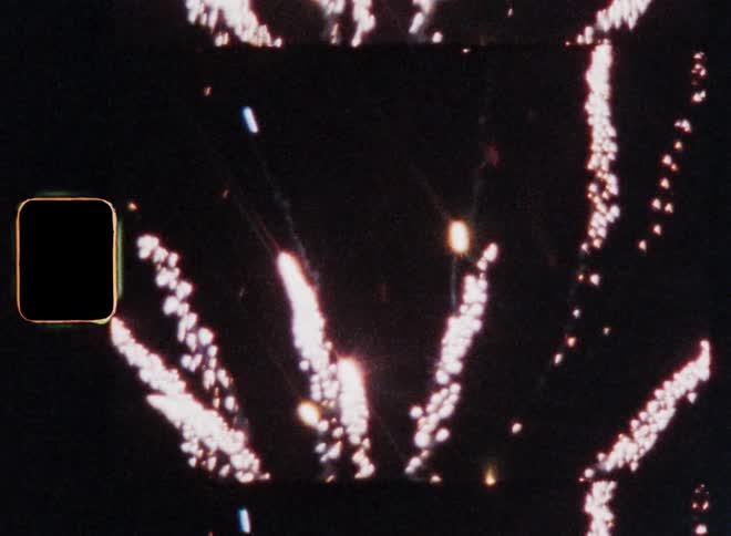 A firework comes out of a box on the ground and explodes in the dark or black night sky causing several sparks caught on vintage film in the 1980s with shaky and dynamic camera movement