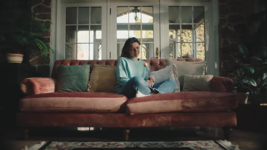 A woman sits on a couch staring at a flower she slowly turns with her hand
