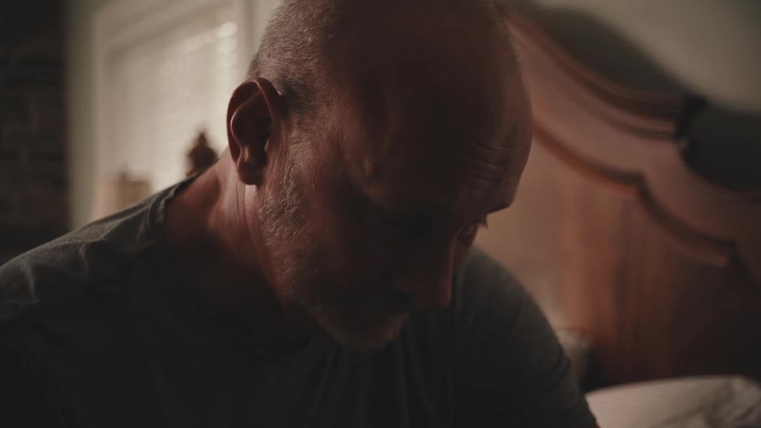 A balding man sits on a bed brooding