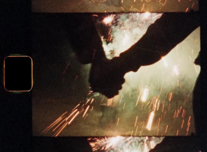 Dynamic retro Super 8 film footage of a man and woman approaching each other grabbing and holding their hands while moving and dancing around expressing joy and excitement in front of a fireworks fountain display