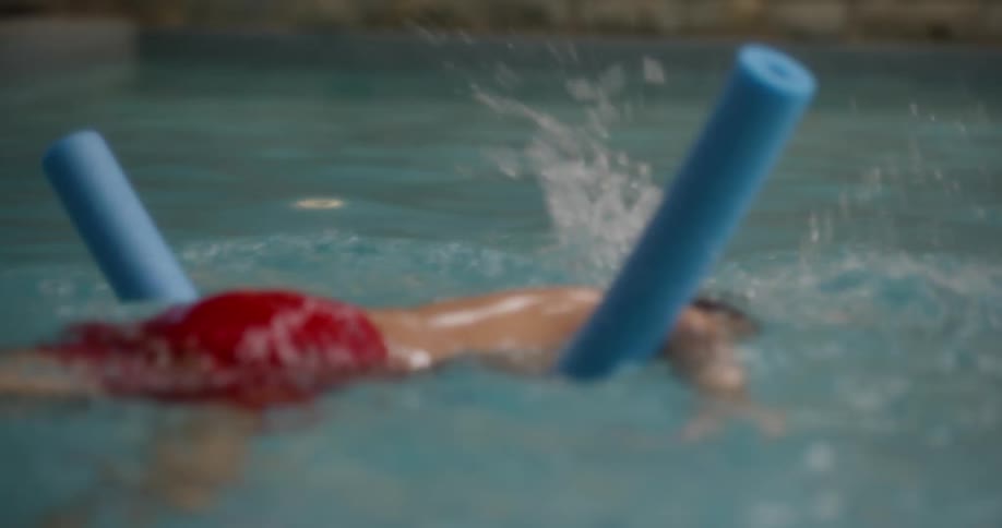 A young boy is leaning on a blue pool noodle to swim