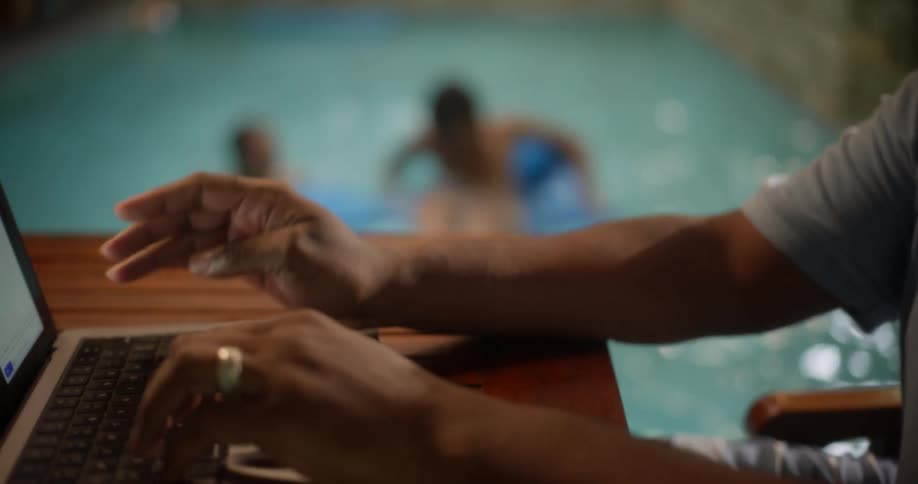 Man sitting concentrated typing on a notebook and in the background two children in the pool