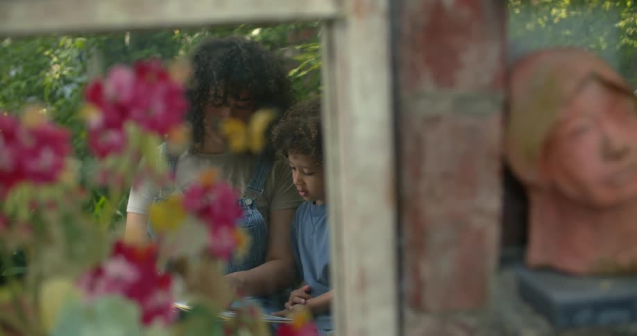 Window POV of a young woman sitting next to a child reading a book in the garden