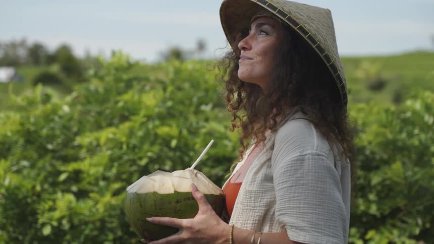 Woman with a hat smiling in a field drinking coconut water