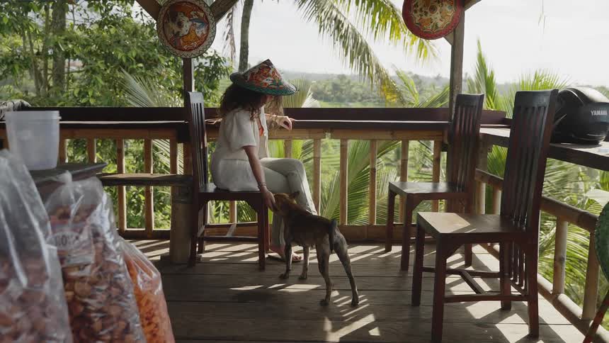 A young woman pets a dog while sitting on an elevated deck in the tropics