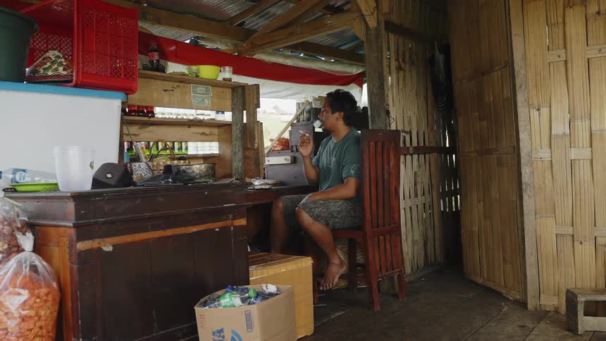 A man sitting at a wooden counter in an open air food stall smoking a cigarette