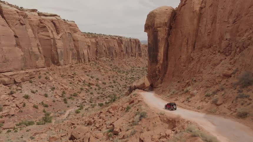 A red off road vehicle driving on a dirt road through a vast desert canyon with steep rocky walls