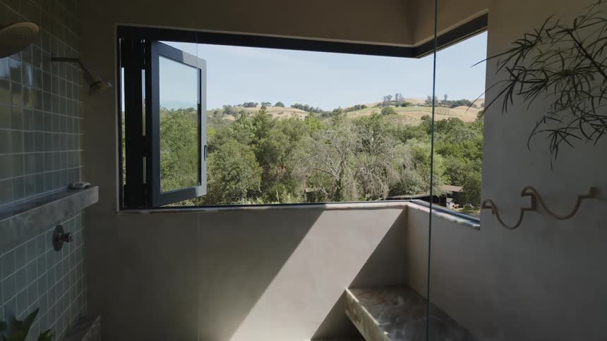 An interior bathroom with grey tiles and large windows overlooking green hills and trees