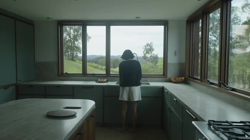Woman walks into a kitchen with a view of the rural landscape and washes produce in the kitchen sink