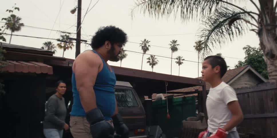 A little boy has his hand on his stomach on the ground in pain and the man next to him lifts him up and they both have boxing gloves on