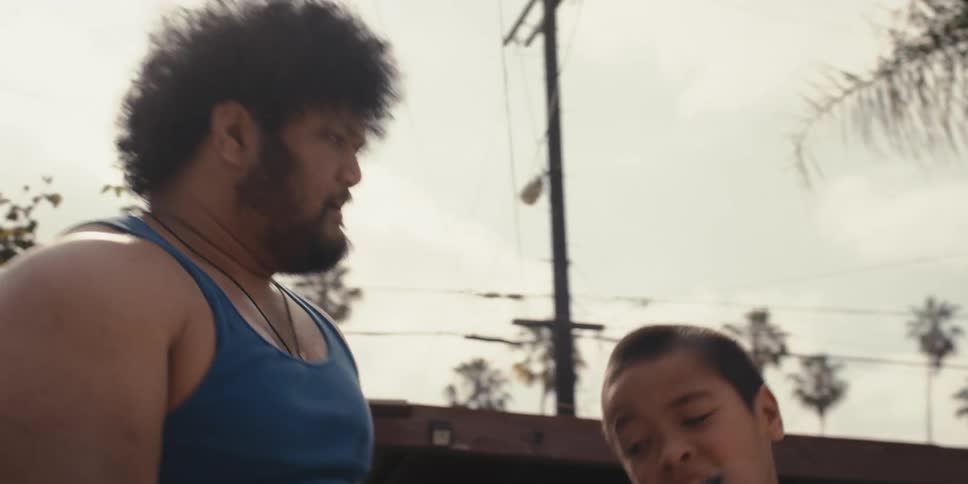 A muscular man wearing a blue undershirt lifts a child for his boxing lessons in the backyard