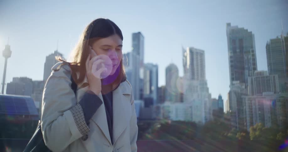 Woman stands outdoors on a sunny day talking on a cellphone with the downtown skyscrapers in the background