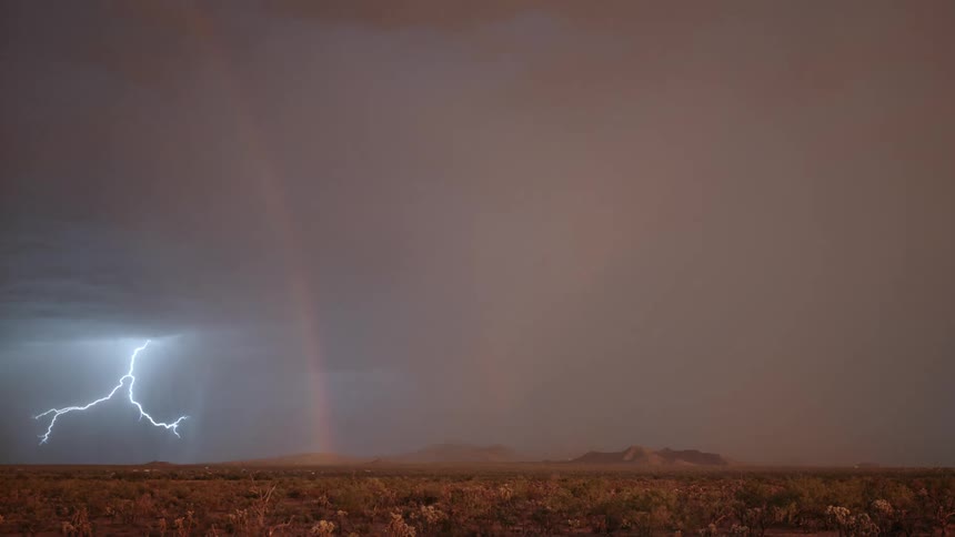 A rainbow hangs down from a stormy sky over mountains and grasslands