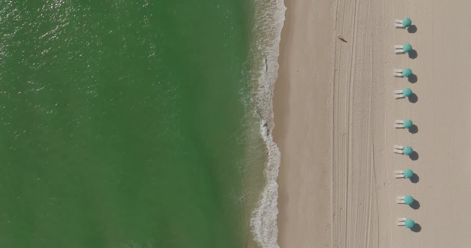 Person walks along the beach between green water and a line of beach furniture