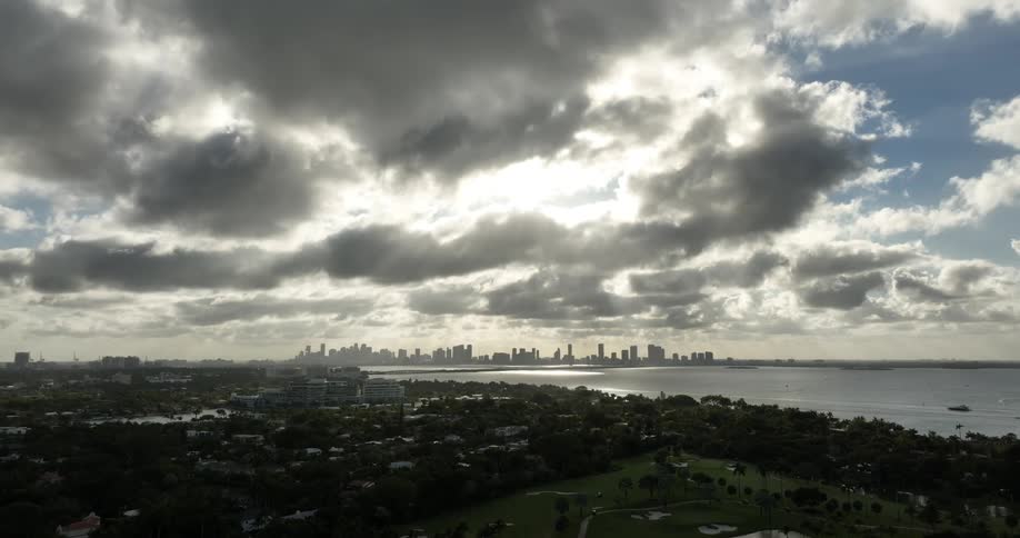 A panoramic view of a city skyline with dramatic cloud cover overlooking a body of water and green areas in the foreground