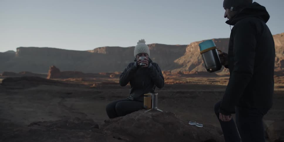 A man and woman have a warm drink as they stand on a peak in a desert landscape