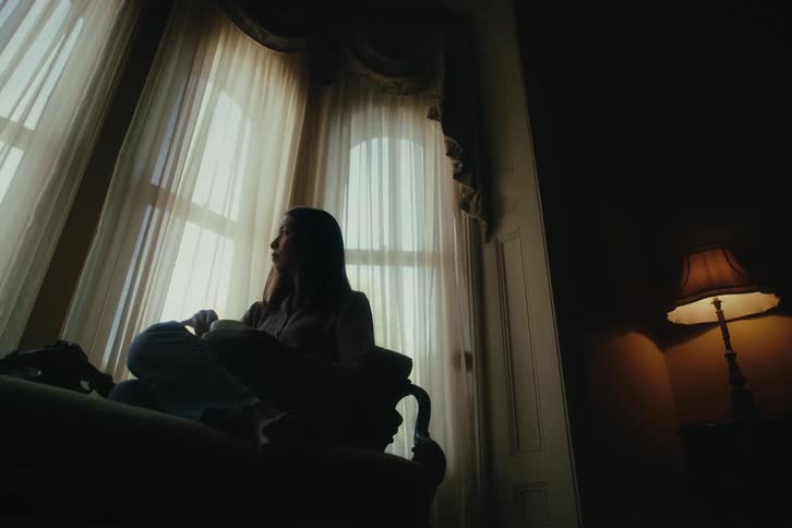 Woman sitting in a dimly lit room looking at a window with a curtain