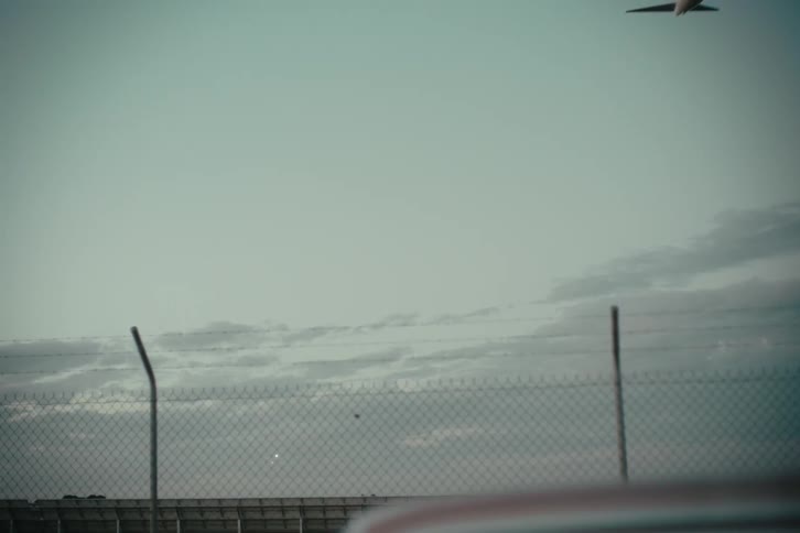 Woman sits in parked car near a fenced field while airplane passes over head