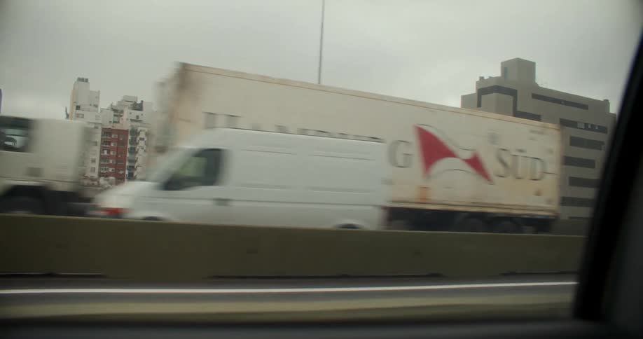 View from the window of a moving vehicle with vehicle traffic on an overpass and buildings in a metropolis on a cloudy day