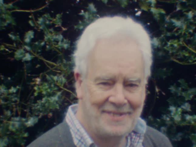 An elderly man in front of a tree takes off his glasses and smiles
