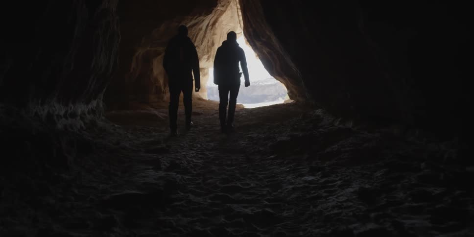 Two people walking inside a dark cave head towards the sunlit opening