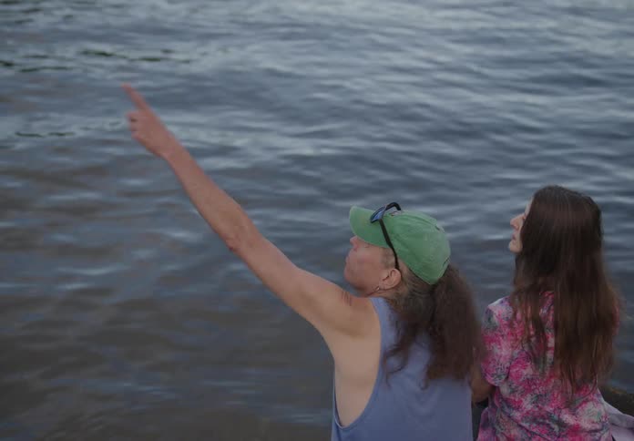 A couple of women sitting on the coast of a lake looking and pointing around