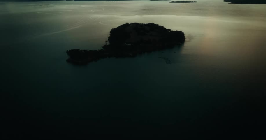 A small dark island sits in the middle of a calm reflective body of water at dusk