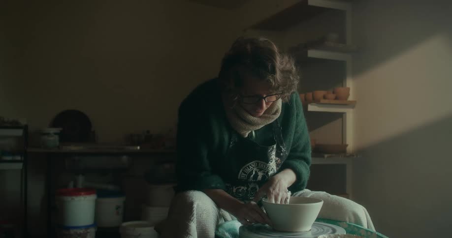 A woman in a green sweater shaping clay on a potters wheel in a softly lit workshop