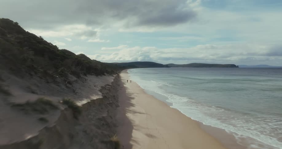 Fast aerial flyover two people walking on the sand along the shoreline as waves come in during the day