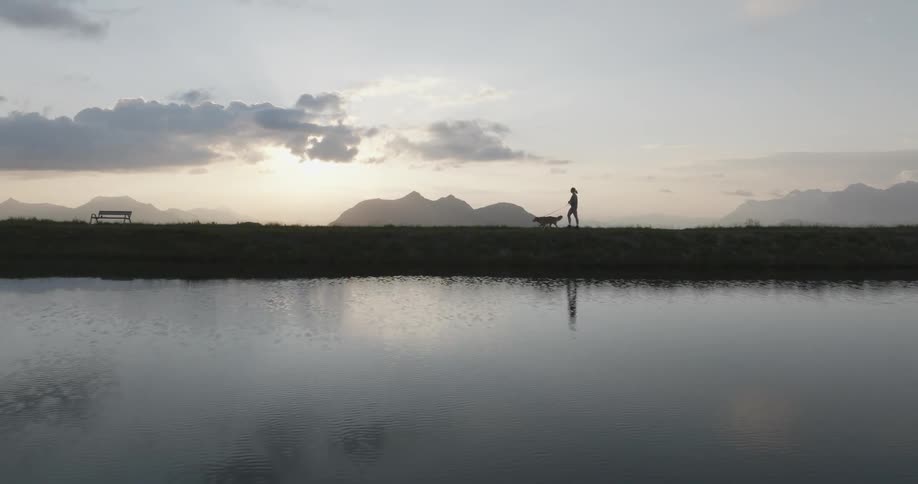 A woman walks her dog beside a lake as the morning sun lights the horizon with fog in the background