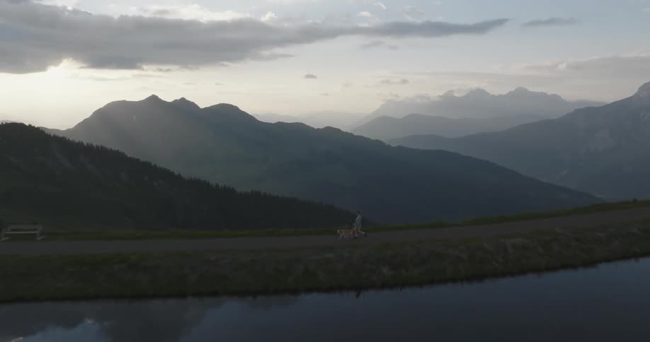 A woman walks her dog beside a clear lake reflecting the sky and clouds with a fog covered mountain range in the background in the morning