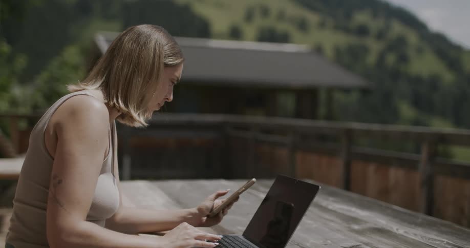 Woman using a laptop and holding a cell phone sitting at a wooden table outdoors with mountains and vegetation in the background