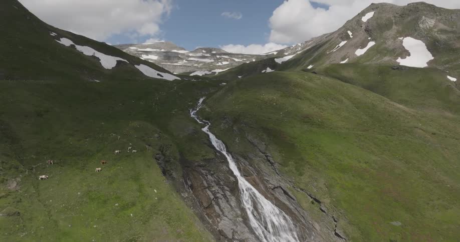 A picturesque waterfall cascades down a green mountainside with snowy peaks in the background