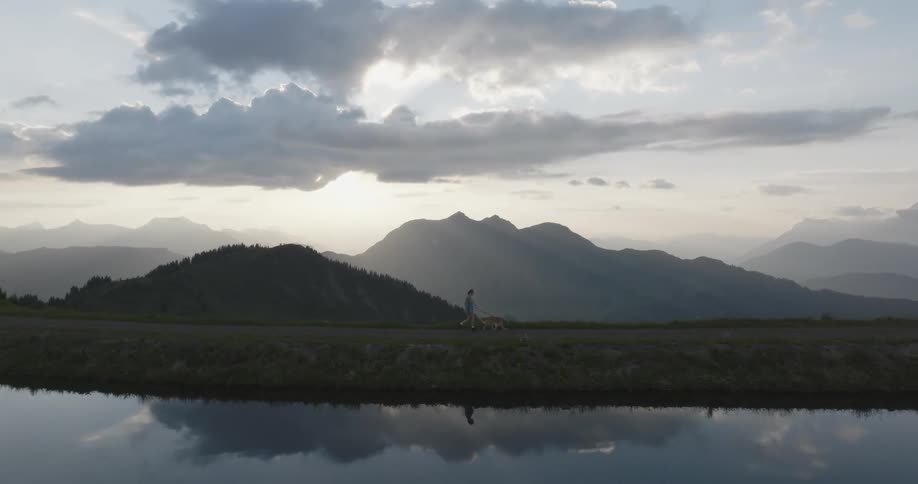 A woman walks her dog next to a reflective body of water with mountains and a cloudy sky in the background