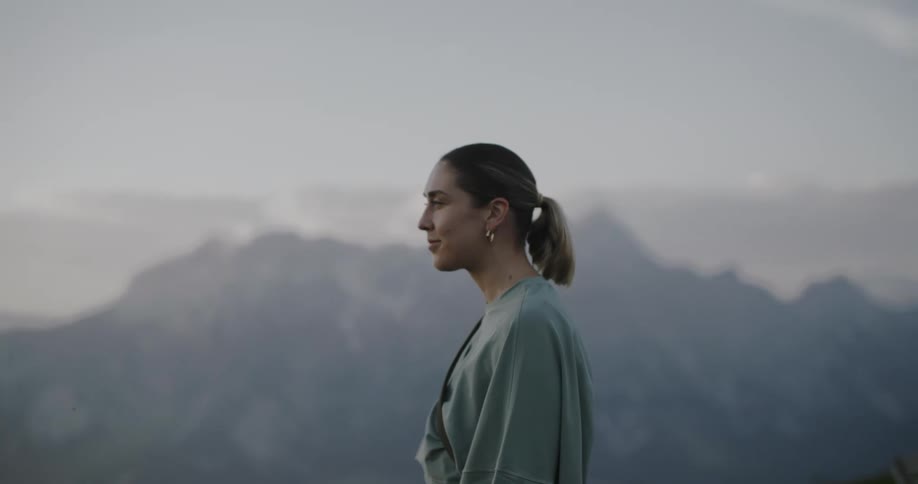 A woman is standing looking in front of her and then looks to the side where there is a mountain range